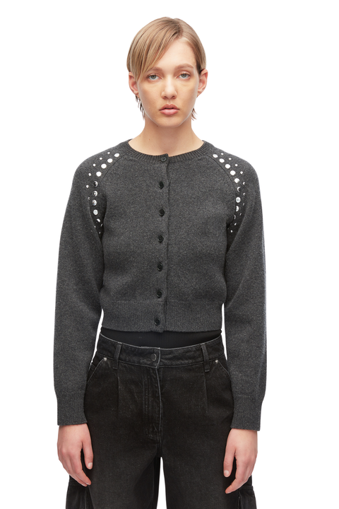 A model wearing a dark gray cropped button-up cardigan with silver stud embellishments on the shoulders, paired with black jeans, against a plain white background.