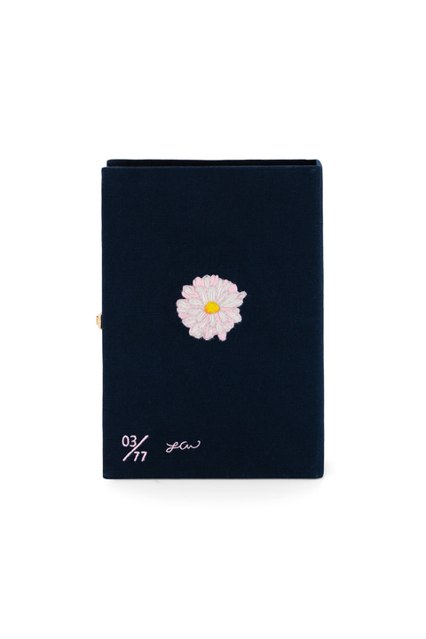 A dark navy blue book clutch with a single light pink daisy embroidered on the front cover, featuring white script text near the bottom left, set against a plain white background.