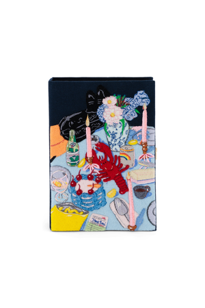 A book clutch featuring a colorful, whimsical illustration of a dinner table scene with a black cat, printed on a dark navy blue cover, shown against a plain white background.