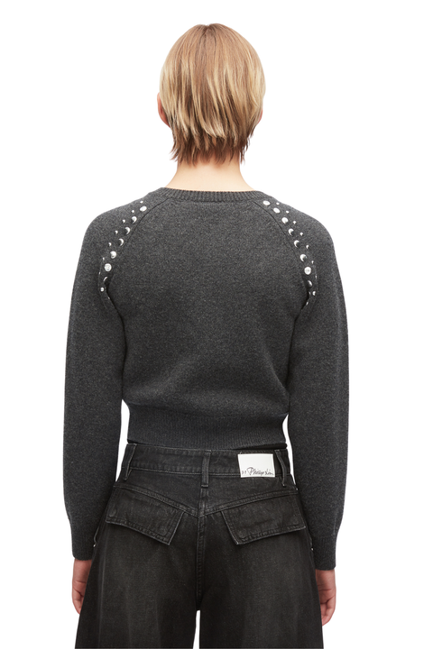 Back view of a model wearing a dark gray knit sweater with silver rhinestone embellishments on the shoulders, paired with dark jeans.
