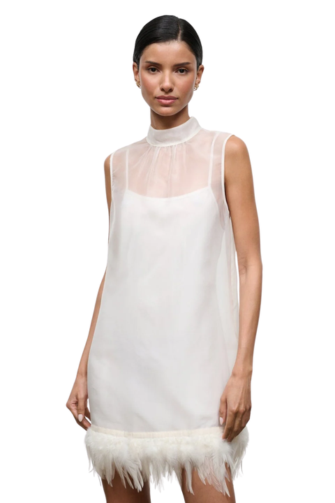 A model wearing a sleeveless white high-neck mini dress with a sheer mesh yoke and a fluffy feather-trimmed hem, set against a plain white background.