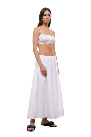 A woman modeling a white two-piece outfit consisting of a cropped camisole top with thin straps and a matching high-waisted maxi skirt, paired with black slide sandals, against a plain white background.