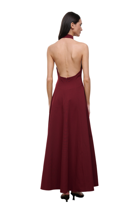 A woman shown from the back wearing a sleeveless, open-back floor-length gown in deep burgundy, paired with black heels, against a plain white background.