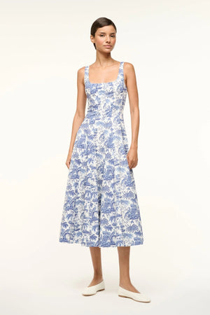 Woman wearing a sleeveless, blue toile midi dress that features a square neck and corset paneling, paired with white flats.