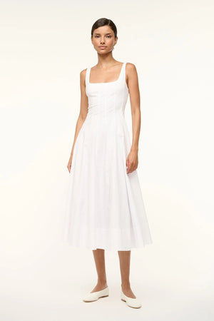 Woman wearing a sleeveless white midi dress that features a square neck, corset-style paneling, and an A-line skirt, paired with white flats.