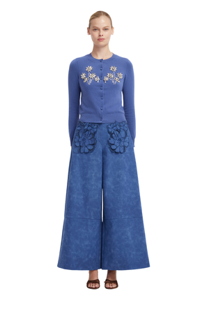 A model stands against a white background wearing a blue long-sleeve button-front top with floral embroidery on the chest and matching wide-leg pants with large floral appliqué details at the hips.