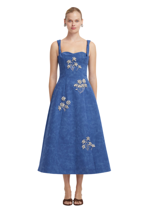 A woman modeling a sleeveless midi dress in a dusty medium blue fabric, featuring white floral embroidery on the bodice and skirt, set against a plain white background.