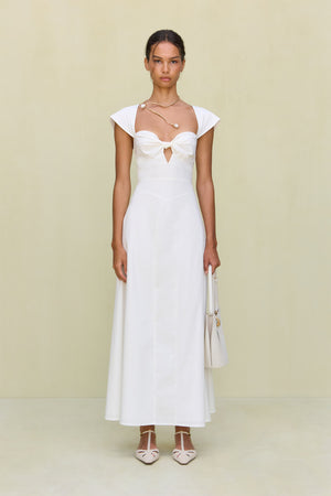 Woman wearing a white maxi poplin dress that features a sweetheart neck, cap sleeves, and a cutout at the bust, paired with white sandals.