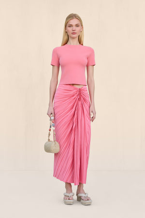 Woman wearing a pink pleated midi skirt that features an asymmetrical waistline and gathered ruching at the hip, paired with a pink matching tee and tan sandals.