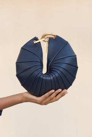 A navy blue leather clutch inspired by a sea shell design with a gold hardware interlocking-clasp closure, held up by a woman's hand.
