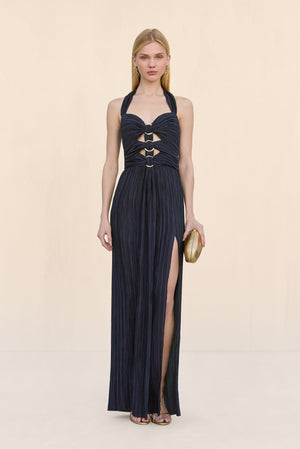 Woman wearing a navy plisse halter gown that features a sweetheart neckline, cutouts at the chest fasten by gold rings, and a high leg slit, paired with gold accessories.