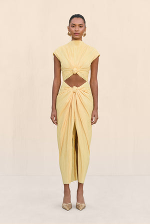 Woman wearing a structured midi dress crafted from a light yellow plisse fabric that features a mock neckline, cap sleeves, cutout at the waist framed by knotted ruching, and a draped center slit, paired with gold heels.