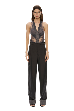 A model stands against a plain white background wearing a sleeveless black halter top with sheer lace panels and high-waisted wide-leg trousers.