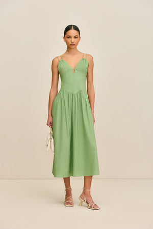 Woman wearing a green, basque waist midi dress that features a double spaghetti strap detail, a deep-V and a small keyhole cutout at the bust, paired with tan heels and a white handbag.