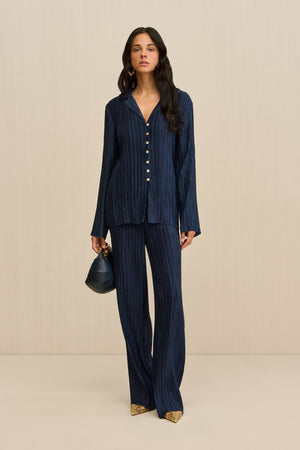 Woman wearing a navy plisse button down shirt that features pearlescent contrast buttons, paired with matching pants and a navy handbag.