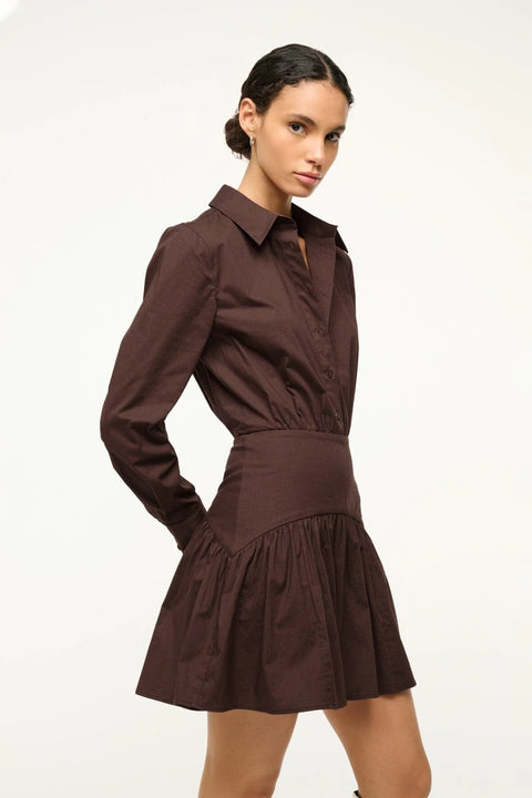 Close up shot of a woman wearing a brown poplin mini dress that features a button down front, long sleeves, and a curved yoke detail at the hip.
