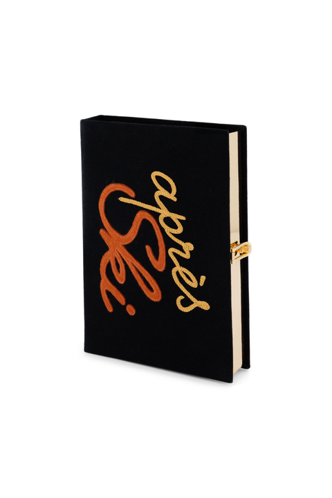 A black book clutch with a gold and orange metallic script cover reading 'Après Ski', featuring a gold clasp closure on the side.