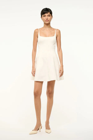 Woman wearing a white mini dress that features a formfitting bodice with a scoop square neck and a flouncy skirt, paired with cream heels.