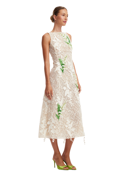 A woman modeling a sleeveless cream lace midi dress with green embroidered leaf motifs, paired with lime green high heels, against a plain white background.