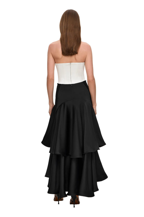 Woman facing away wearing a strapless white top with zipper and black ruffled maxi skirt on a white background