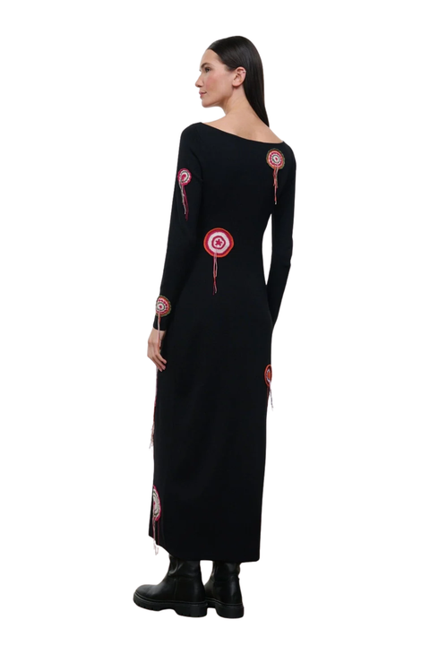 A woman modeling a long black dress with red and white circular target motifs on the shoulders, sleeves, and hem, paired with black ankle boots, shown from a side-back angle against a plain white background.
