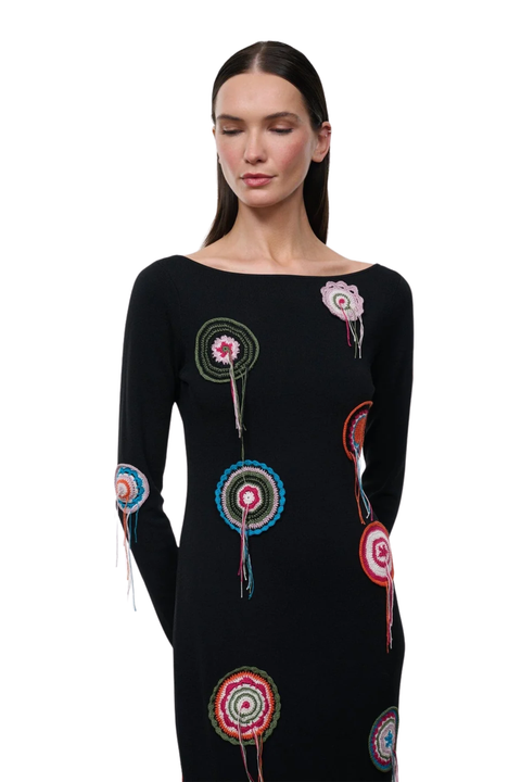 A woman modeling a black long-sleeve knit dress with colorful circular floral embroidery and fringe details, set against a plain white background.