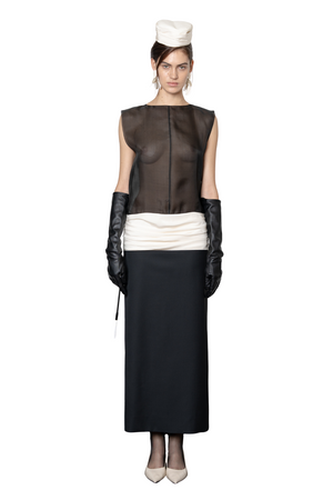 A model stands against a white background wearing a black and white long dress consisting of a sheer black boat neck sleeveless top, a white gathered drop waistband, a long black skirt, black gloves, and a white hat.