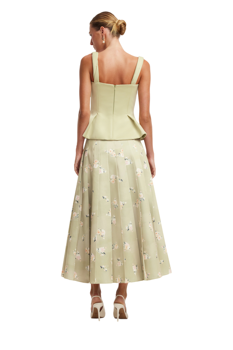 A woman shown from the back wearing a light sage green sleeveless dress with a structured peplum waist and a full skirt featuring a delicate floral print, paired with beige high heels against a plain white background.