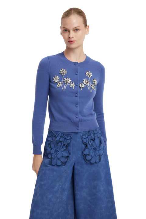 A model wearing a blue button-front cardigan with white floral embroidery and matching wide-leg blue pants with large floral applique on the front pockets, set against a plain white background.