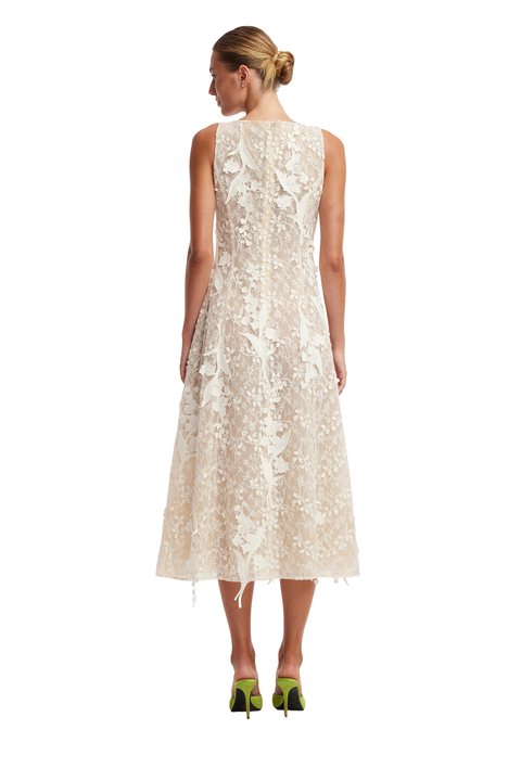 A woman viewed from the back wearing a sleeveless cream lace midi dress with a fitted bodice and flared skirt, paired with bright green heels, against a plain white studio background.