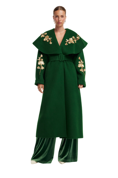 A model wearing a dark green long coat with a wide collar, matching belt, and floral embroidery on the shoulders and sleeves, paired with matching green wide-leg trousers, against a plain white background.