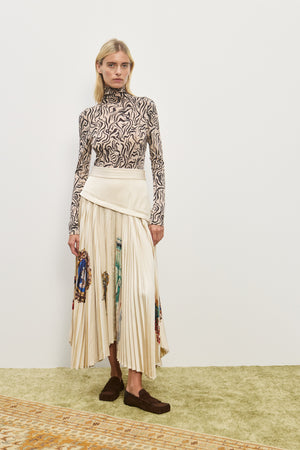 Woman wearing a cream midi skirt with an asymmetrical corseted waist and a pleated skirt with an asymmetrical hem line and colorful icons printed on the skirt, paired with a printed long sleeve top and brown loafers.