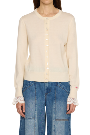 A model wearing a cream-colored long-sleeve knit cardigan with lace ruffle cuffs and a button-up front, paired with blue denim jeans against a plain white background.