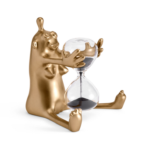 Small gold figurine sitting while holding glass sand timer in hands on white background.