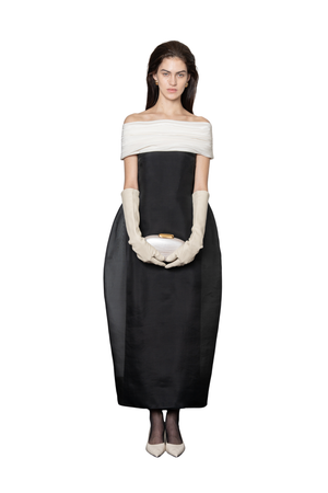 A model stands against a white background wearing a black off-the-shoulder midi dress with a wide cream band across the upper chest, cream elbow-length gloves, and holding a small cream clutch.