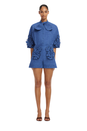 A model stands against a white background wearing a blue short matching set with a large ruffled collar, elbow-length sleeves with 3D floral applique, and matching floral details on the front pockets.