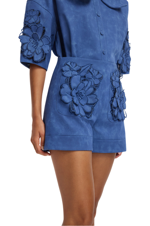 A model wearing a blue short-sleeve matching set with large 3D floral appliques on the front pockets and sleeve cuffs, shown against a plain white background.
