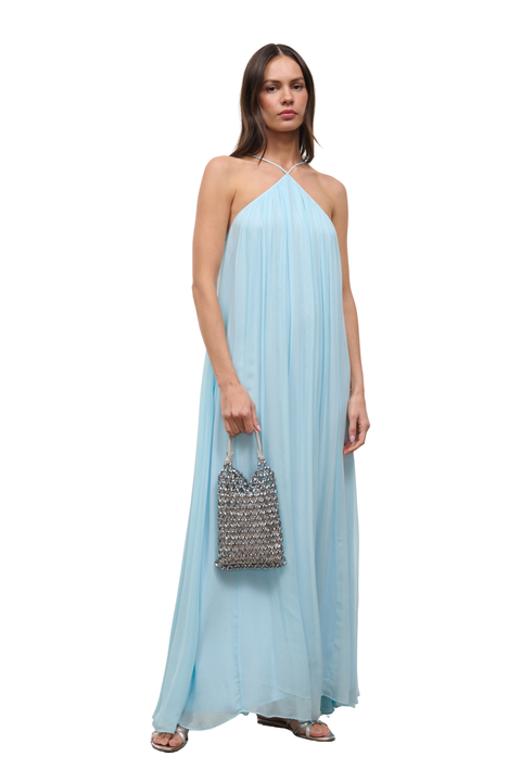 A woman modeling a light blue halter-neck maxi dress with a flowing chiffon-like fabric, holding a small woven metallic handbag against a plain white background.