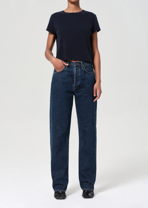 Person wearing a navy t-shirt and blue jeans on a white background with hand in front pocket.