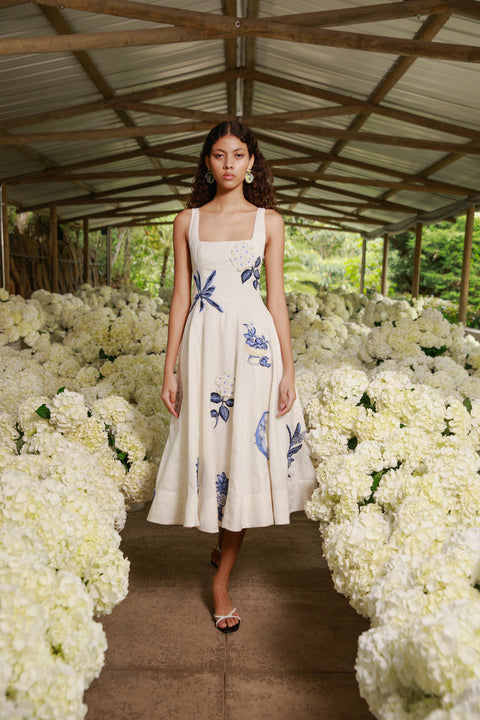 Model wears white linen midi dress with blue embroidery on runway lined with white flowers.