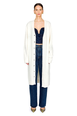 Woman wearing a long white cardigan over a dark top and jeans and pearl necklace on a white background