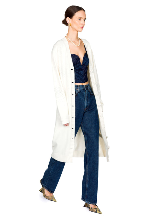 Woman walking in  a long white cardigan with crystal embellishments over a navy top and jeans on a white background