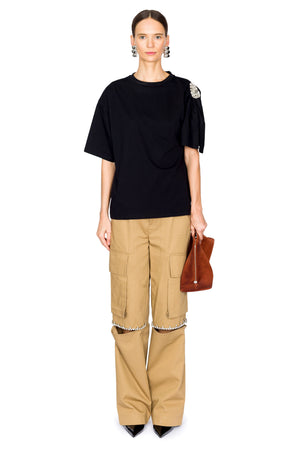 Person wearing a black short sleeve top with crystal embellishment on one shoulder and beige cargo pants with a white background