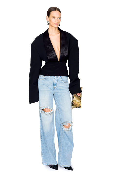 Woman holding snakeskin clutch wearing a black deep v-neck long sleeve top and light blue wide leg jeans on a white background