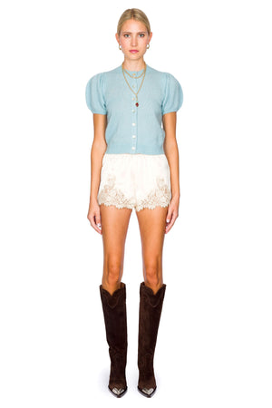 Woman wearing a light blue short sleeve cardigan and white lace shorts, and brown knee-high boots on a white background