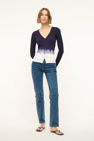 Woman wearing a fitted, rib-knit cardigan that features a deep-V and a navy to white tie dye effect, paired with jeans and sandals.