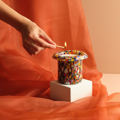 Hand reaching out to light multicolored glass candle with orange tulle background.