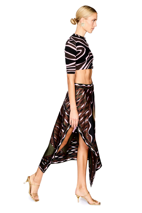 A model is shown in profile wearing a matching two-piece outfit consisting of a short-sleeve crop top sweater and a high-low skirt, both featuring a wavy abstract pattern in black, brown, and pink. She is wearing gold strappy high-heeled sandals against a plain white background.
