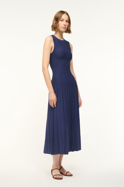 Side angle of a woman wearing a navy knit midi dress that features a high neckline and flattering ribbing in the torso with a pleated skirt.