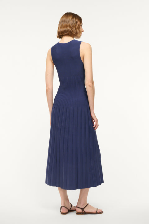 back angle of a woman wearing a navy knit midi dress that features a high neckline and flattering ribbing in the torso with a pleated skirt.